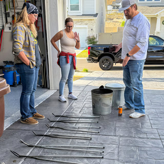 Three people stand on a driveway beside metal buckets and a row of long tongs laid out on the ground during a raku firing with The Clay Hole, a pottery studio in Draper, Utah