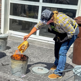 A person in a bandana and plaid shirt lowers a brown paper bag toward a metal bucket of flames during a raku firing with The Clay Hole, a pottery studio in Draper, Utah