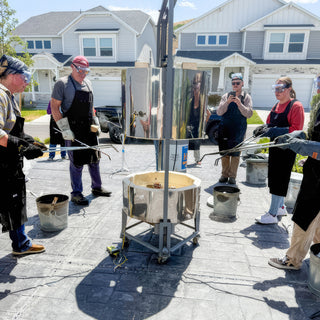 A circle of people in goggles and aprons stand around a metal kiln on wheels, holding long tongs during a raku firing with The Clay Hole, a pottery studio in Draper, Utah