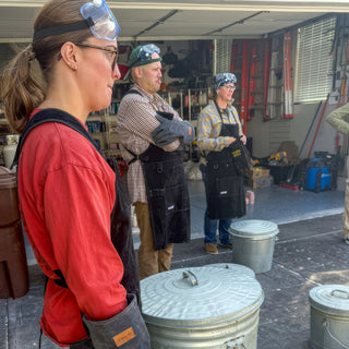 A person in a red shirt and black apron stands in profile beside lidded metal buckets, two more behind during a raku firing with The Clay Hole, a pottery studio in Draper, Utah