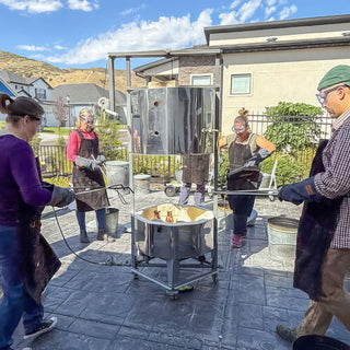 A group of potters in goggles, gloves and aprons stand around a shiny metal kiln with its lid raised outdoors during a raku firing with The Clay Hole, a pottery studio in Draper, Utah