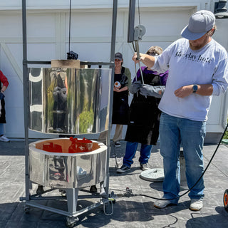 A person in a cap raises the lid of a shiny metal kiln on a stand with red pieces inside during a raku firing with The Clay Hole, a pottery studio in Draper, Utah