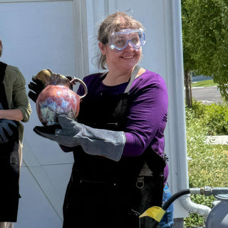 A smiling potter in goggles and a long glove holds a round pitcher with a shiny copper and purple glaze during a raku firing with The Clay Hole, a pottery studio in Draper, Utah