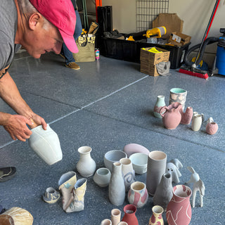 A person in a pink cap holds a white vase above small pastel vases and bowls on a gray floor during a raku firing with The Clay Hole, a pottery studio in Draper, Utah