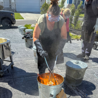 A potter in goggles, gloves and a black apron leans over a smoking bucket of flames with tongs during a raku firing with The Clay Hole, a pottery studio in Draper, Utah