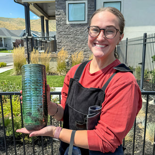 A smiling potter in a red shirt and black apron holds a tall ribbed cylinder vase with shiny teal glaze during a raku firing with The Clay Hole, a pottery studio in Draper, Utah