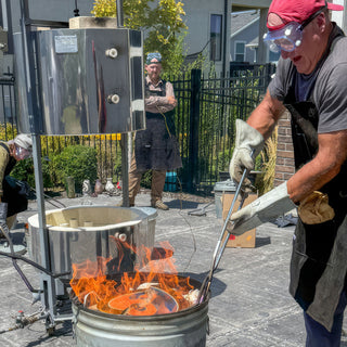 A person in a red cap and goggles reaches long tongs into a bucket of flames beside a metal kiln during a raku firing with The Clay Hole, a pottery studio in Draper, Utah