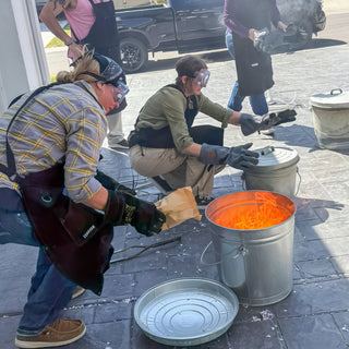 Two potters crouch beside metal buckets, one holding a brown paper bag over a bucket glowing orange inside during a raku firing with The Clay Hole, a pottery studio in Draper, Utah