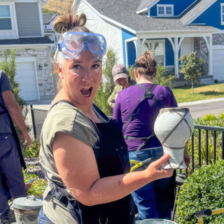 A potter with goggles on their head holds up a white vase with gray lines crossing its surface outdoors during a raku firing with The Clay Hole, a pottery studio in Draper, Utah