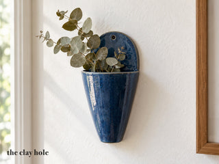 A deep blue stoneware wall pocket vase hanging on a wall with eucalyptus, made from The Clay Hole wall pocket paper template